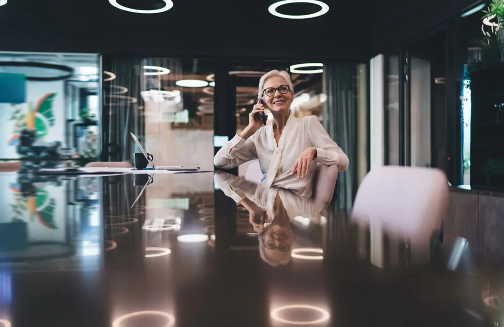 Smiling businesswoman talking on the phone in a modern conference room with a reflective table and circular ceiling lights, representing procurement transformation services.